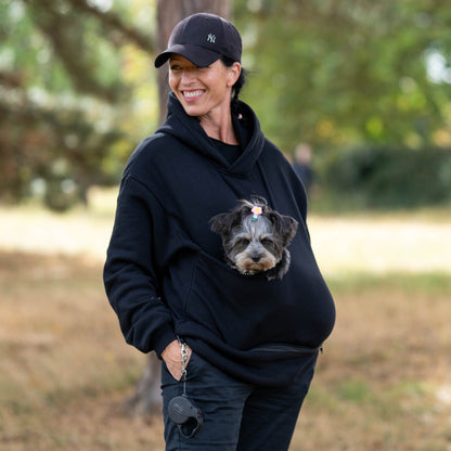 Woman in a black dog hoodie carrier and cap holding a small dog in a pouch. 