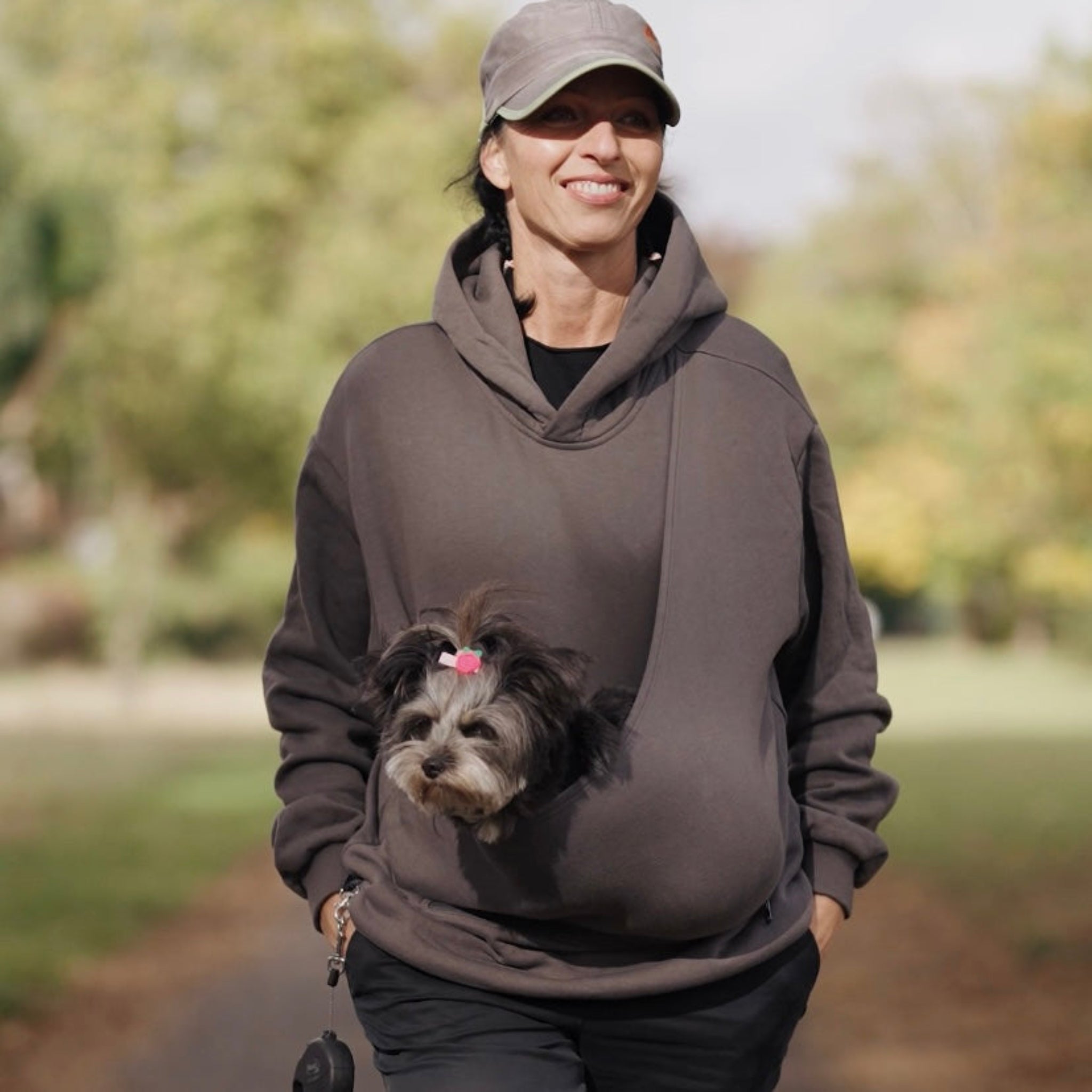 Woman wearing a brown dog hoodie carrier with a dog peeking out, standing outdoors.